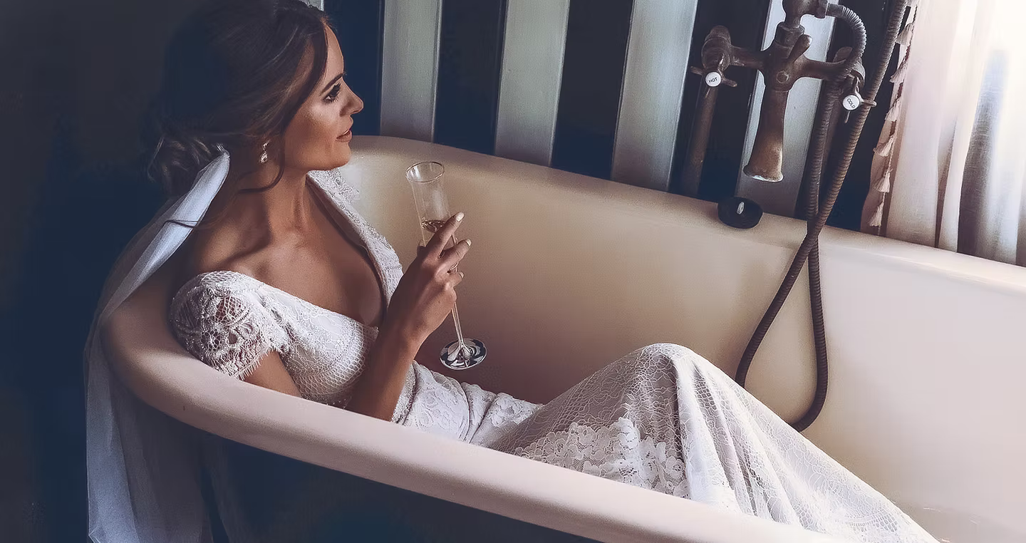 Bride in lace gown lounging in vintage clawfoot tub with champagne during bridal prep